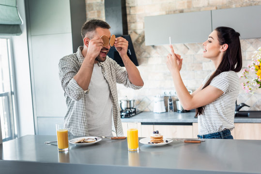 woman taking picture of husband playing with breakfast at counter in kitchen
