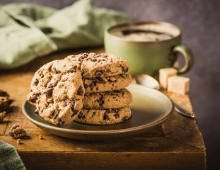 Stack of chocolate chip cookies on green plate with cup of coffee on old wooden table. Selective focus. Retro style toned.