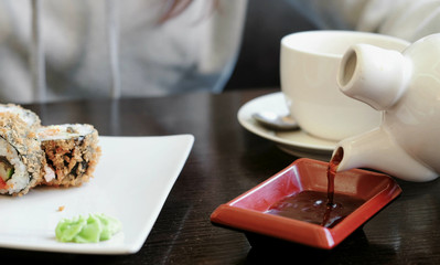 Woman's hand pours soy sauce in small rectangular plate, rolls and cup of tea on background.