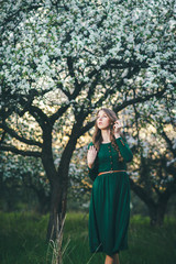 Young beautiful woman in green dress posing in the blooming garden at the sunset