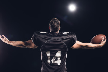rear view of american football player with ball looking up with outstretched hands under spotlight on black