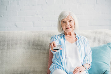 portrait of smiling senior woman with remote control watching tv on sofa at home