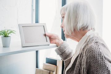 side view of senior lady looking at empty photo frame on bookshelf at home