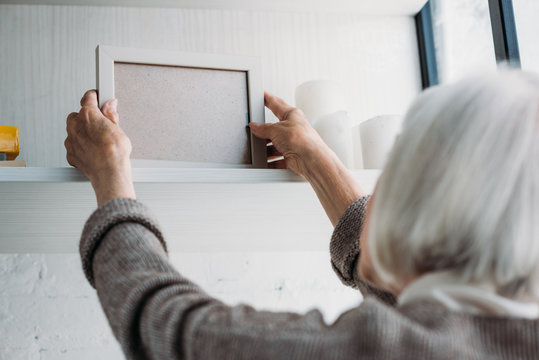 partial view of senior lady taking empty photo frame from bookshelf at home