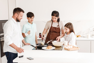 Joyful family woman and man with happy kids 8-10 having fun, and cooking pancakes together at home