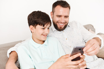 Image of caucasian man 30s and son sitting together on sofa at home, and using black smartphone