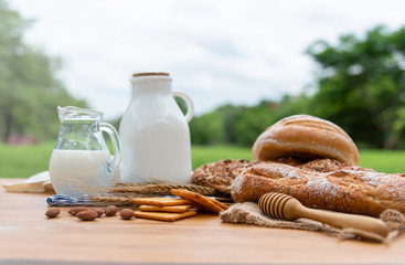 Photo of bread, bun, milk, almonds and fresh milk on wooden table with blurred park background. Breakfast preparation, daily product.