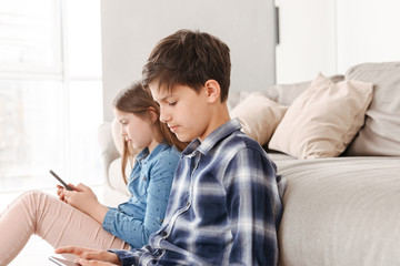 Fototapeta premium Image of young siblings girl and boy 8-10 sitting on floor near sofa at home, and both using smartphone