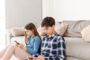 Portrait of european siblings girl and boy 8-10 sitting on floor near sofa at home, and both using...