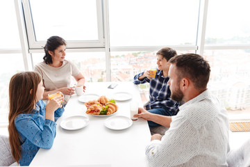 Photo of attractive people parents with children 8-10, having breakfast together in bright kitchen at home and eating croissant sandwiches