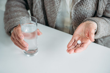 partial view of senior woman with pill and glass of water in hands sitting at table alone at home