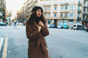 Fototapeta premium Business woman wearing light brown coat and black beret standing beside road while waiting for the taxi to go home after hard working day. Beautiful caucasian female with dark curly hair walking city.