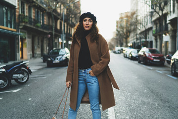 Obraz premium Beautiful model look woman with long dark curly hair wearing stylish clothes standing in the middle of the road on a blurred street background. Stylish blogger walking the city in the evening.