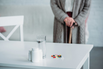 selective focus of senior woman with walking stick standing in room with medicines and glass of water on tabletop at home
