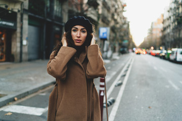 Beautiful caucaisan female with long dark curly hair wearing light brown coat adjusting a black beret on her head while standing on a street while waiting for the taxi to go to the party with a friend