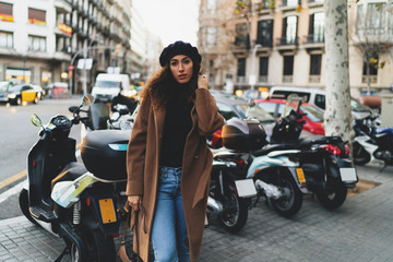 Portrait of a pensive caucasian female with dark curly hair wearing coat, jeans, sweater and a beret standing outdoors with a bag in her hand on a blurred street background with parked motorcycles.