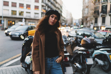 Obraz premium Charming hipster girl in a trendy coat and a beret looking at the camera while standing on blurred street road background. Student girl with dark curly hair enjoying free time in the evening outdoors.