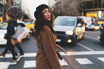 Portrait of a charming caucasian woman with beautiful long dark curly hair wearing coat and a beret looking back at the camera while crossing the street on a blurred urban background.