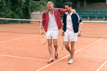 cheerful sportive friends with wooden rackets hugging and walking on tennis court