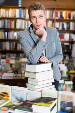 Pensive young man posing on stack of books in bookstore
