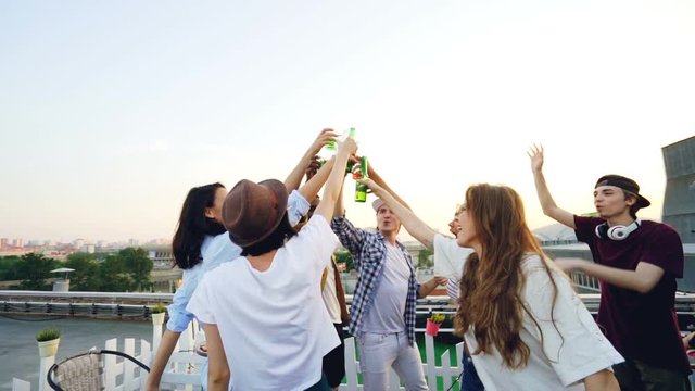 Slow motion of excited friends dancing with drinks then clinking bottles and drinking at open-air party on rooftop with deejay using equipment to mix music.