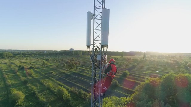aerial view of telecommunications tower, technician in a safety vest and hard hat uses mobile phone on top of cellular antenna