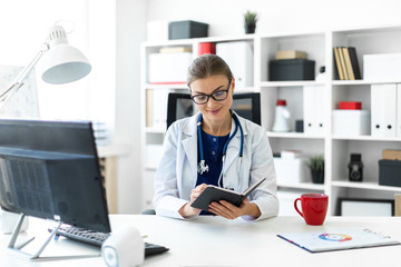 A young girl in a white robe sits at a table in the office and holds a pen and a notebook in her...