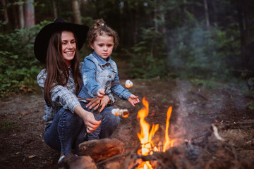 Mother and daughter fry marshmallow on  campfire in forest. Young family traveling active adventure...