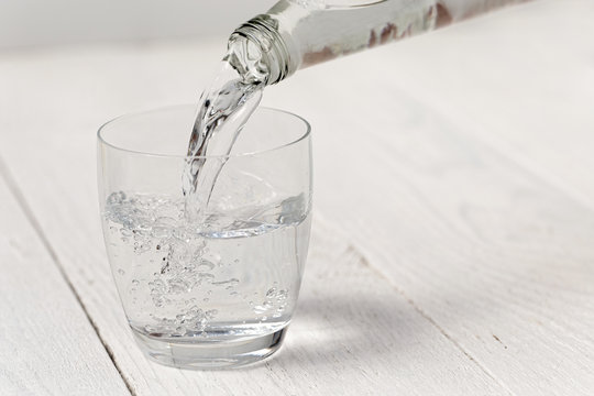 Pouring water from a glass bottle into a glass. White wood background.