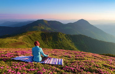 Fototapeta premium On the lawn among the rhododendron flowers the yoga girl is sitting in the pose on the colorful rug. Meditation. The landscape with the mountains in the fog. Nice summer scenery.