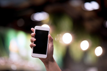 Mockup image of a woman holding and displaying a white mobile phone with a black screen with a blank screen on a table in a modern cafe. Ideas for working with communication tools.