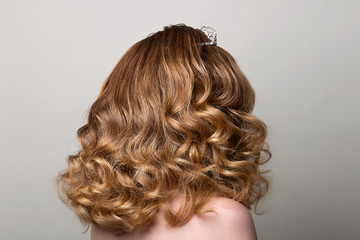 Female hairstyle long curls on the head of the brown-haired woman looking back at the gray background turning the head to the right.