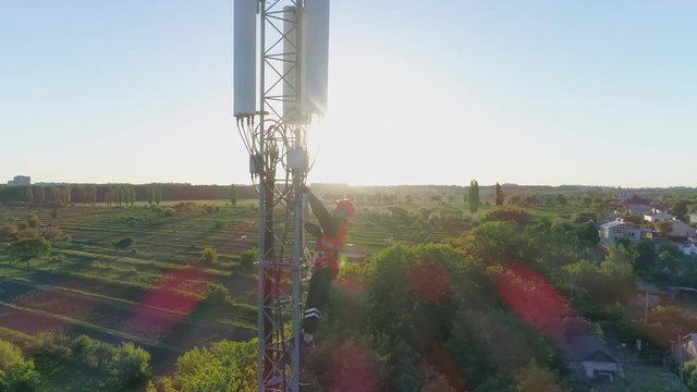 engineer in a safety vest and hard hat climbs high on cellular antenna, drone view of telecommunication tower