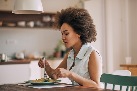 Beautiful mixed race woman eating pasta for dinner while sitting at kitchen table.