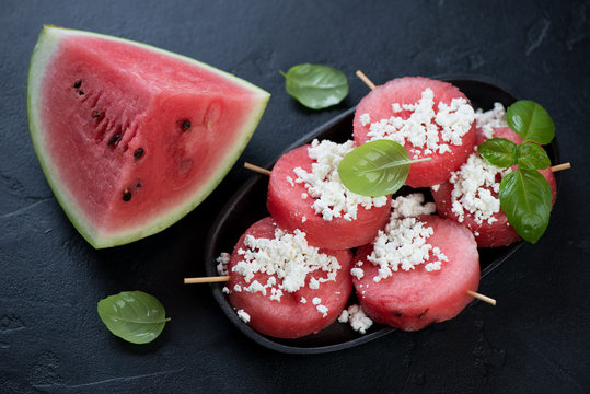 Cold summer dessert made of round fresh watermelon slices on wooden skewers with cottage cheese, studio shot on a black stone background