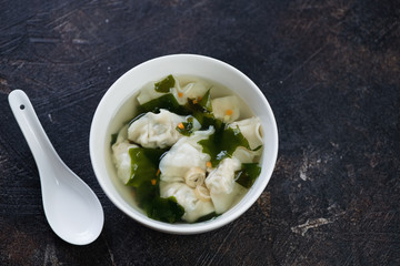 White bowl of wonton soup over dark brown stone background, high angle view with space