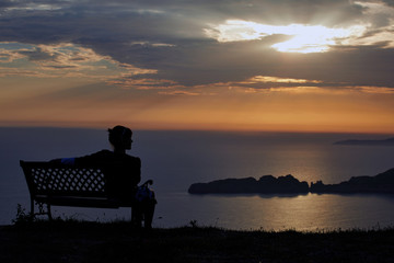 Fototapeta na wymiar Young woman sits on a bench on a sunset background over the sea and mountains