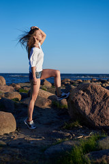 The girl stands on a large stone against the backdrop of rocks and the sea and enjoys the sunset
