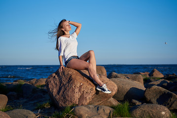 The girl sits on a large stone against the backdrop of rocks and the sea and enjoys the sunset
