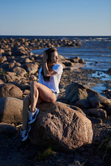 The girl sits on a large stone against the backdrop of rocks and the sea and enjoys the sunset