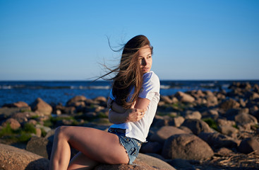 Fototapeta na wymiar The girl sits on a large stone against the backdrop of rocks and the sea and enjoys the sunset