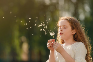 Cute curly girl blowing dandelion in sunset light.