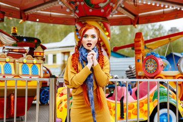 young cheerful red-haired girl posing on the background of the carousel in the amusement Park. Portrait of a woman with bright makeup and long red hair braided in braids. She poses, has fun and plays.