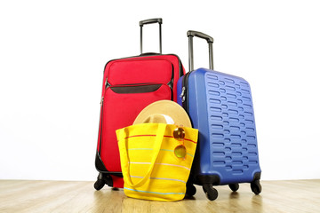 Red textile suitcase & blue hard shell luggage, extended telescopic handle, straw hat, yellow beach bag, mirrored sunglasses, white wall background. Couples retreat trip concept. Close up, copy space.