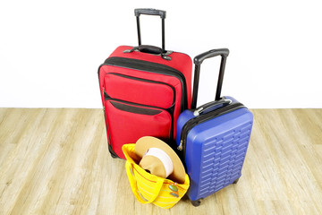 Red textile suitcase & blue hard shell luggage, extended telescopic handle, straw hat, yellow beach bag, mirrored sunglasses, white wall background. Couples retreat trip concept. Close up, copy space.