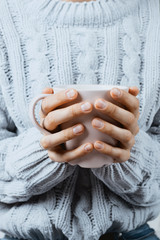 Closeup of woman's hands in a blue sweater holding pink mug with warm beverage, selective focus