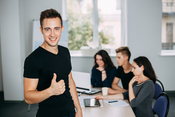 Cheerful confident young man gesturing thumb up with his colleagues working behind him. friendly atmosphere, cohesive team, communication and comradery. 
