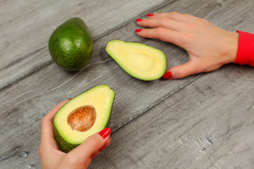 Top view - woman hand with red nails, holding avocado cut in half, with other whole green pear in back, on gray wood table