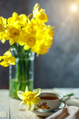 a bouquet of yellow daffodils is in a vase and a Cup of tea on the table in the morning still life