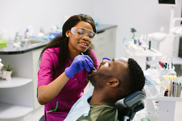 Young african american man lying in chair receiving dental treatment with mouth open, dentist hands wearing gloves holding tools working on patients teeth - Powered by Adobe
